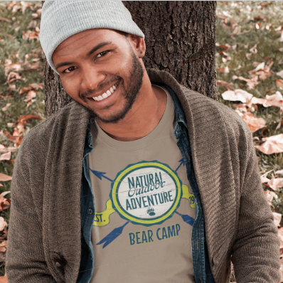 Gay Man sitting outdoors with a book, wearing a gay bear camp t-shirt with 'Natural Adventure Bear Camp' design.